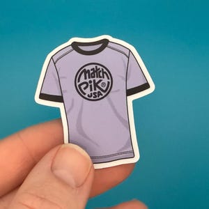May include: A sticker of a light purple t-shirt with black trim and the words "Match Pik USA" in a circular design on the front. The sticker is held by a hand against a blue background.