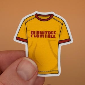 May include: A yellow sticker in the shape of a t-shirt with the word "PLUMTREE" in maroon block letters across the chest. The shirt has maroon trim on the collar and sleeves.