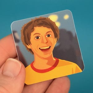 May include: A square sticker featuring a smiling cartoon portrait of a person with brown hair, wearing a yellow shirt with a red collar. The background is a blurred blue and gray. The sticker is held by a hand.
