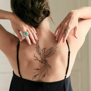 May include: A black line art tattoo of a phoenix bird with outstretched wings, perched on a branch, and with long tail feathers. The tattoo is on a person's back, with their arms raised. The person is wearing a black top.
