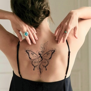 May include: A black butterfly tattoo design on the back. The tattoo features a woman's silhouette within the butterfly wings, with intricate details and swirling patterns. The person is wearing a black top.
