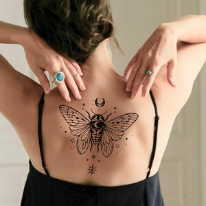 May include: A black temporary tattoo of a cicada with a crescent moon and stars design. The tattoo is on a person's back, with their arms raised. The design is detailed and intricate, with a focus on the insect's wings and body.