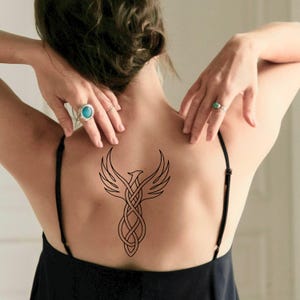 May include: A black temporary tattoo of a phoenix design on a person's back. The phoenix has outstretched wings and a stylized body with intertwined lines. The person is wearing a black top.