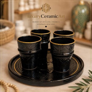 Può includere: Un set di quattro tazze in ceramica nera con bordi dorati e intricati disegni in rilievo, presentate su un vassoio nero abbinato con un bordo dorato. L'immagine include il testo "luxury CeramicArt" e "Moroccan artisanal ceramics".