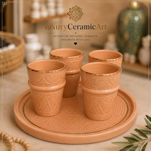 Può includere: Quattro bicchieri da tè in ceramica color terracotta con bordi dorati e motivi in rilievo, presentati su un vassoio rotondo abbinato. Il set fa parte di una collezione di ceramiche artigianali marocchine. Il testo sull'immagine dice "luxury CeramicArt".