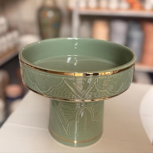 May include: A sage green ceramic pedestal bowl featuring a wide, circular basin and a sturdy base. The exterior is decorated with embossed geometric patterns and delicate floral motifs, accented by a thin, reflective gold rim along the top edge.