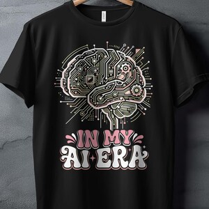 May include: A black short-sleeve t-shirt featuring a graphic of a human brain designed with intricate circuit board patterns and gears. Below the brain, the text reads IN MY AI ERA in a bold, retro-inspired font with pink and white coloring.