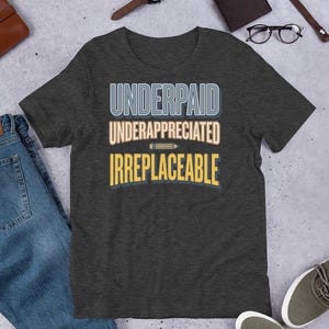 May include: Dark gray t-shirt with the words "UNDERPAID UNDERAPPRECIATED IRREPLACEABLE" in a layered design. The text is in white, light blue, and yellow. The shirt is displayed on a flat surface with jeans, shoes, and accessories.