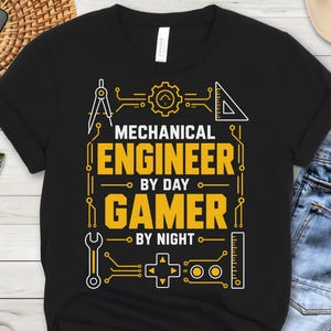 May include: A black short-sleeved t-shirt featuring a yellow and white graphic print with the text "MECHANICAL ENGINEER BY DAY GAMER BY NIGHT". The design includes icons of a gear, a compass, a triangle ruler, a wrench, a game controller, and a ruler.