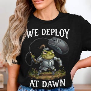 May include: Black t-shirt with the text "WE DEPLOY AT DAWN". The graphic features a cartoon frog in armor holding a whip attached to a computer mouse. The frog is standing on a patch of grass.