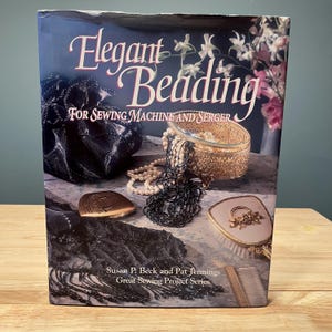 May include: A book titled Elegant Beading For Sewing Machine and Serger by Susan P. Beck and Pat Jennings. The cover features a dark fabric pouch, a gold filigree box with white and black beads, a gold hand mirror, and a comb on a marble surface.