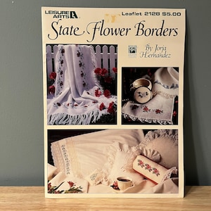 May include: A cross-stitch pattern booklet titled "State Flower Borders" by Jorja Hernandez. The booklet features three images of cross-stitch projects, including a white blanket with red flower borders, a white pillow with red flower borders, and a white napkin with red flower borders. Leaflet 2128 $5.00.