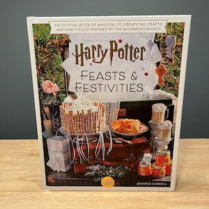 May include: A hardcover book titled Harry Potter Feasts & Festivities, featuring an official guide to magical celebrations, crafts, and party food. The cover displays a cake, pastries, and jars arranged on a vintage brown suitcase.