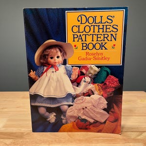 May include: A book titled "Dolls' Clothes Pattern Book" by Roselyn Gadia-Smitley. The cover features a doll wearing a blue and white checkered dress and a straw hat. Various doll clothes are displayed on the cover.