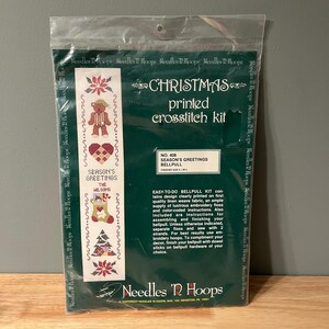 May include: A sealed plastic package containing a Christmas printed cross-stitch kit. The kit features a design with a teddy bear, heart, stocking, and Christmas tree. The text "Season's Greetings" is visible.