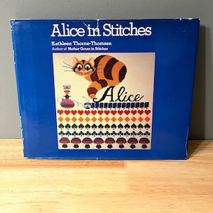 May include: A blue book with the title "Alice in Stitches" by Kathleen Thorne-Thomsen. The cover features a colorful embroidered image of a cat with the name "Alice" in a stylized font.