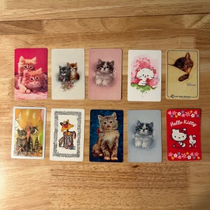 K&ouml;nnte beinhalten: Ein Set aus zehn Vintage-Sammelkarten mit verschiedenen Illustrationen von Katzen. Die Karten sind auf wei&szlig;em Hintergrund mit bunten Bildern von Katzen in verschiedenen Posen und Umgebungen gedruckt. Die Karten enthalten Text wie "Hello Kitty" und "Cherrie".