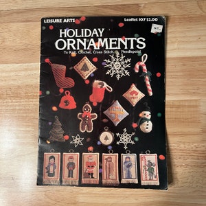 May include: A black and white booklet titled "Holiday Ornaments" with a colorful illustration of various holiday ornaments. The ornaments include a stocking, a snowman, a gingerbread man, a candy cane, a star, a tree, and a bell. The booklet is from Leisure Arts and is Leaflet 107. The text on the cover reads "To Knit, Crochet, Cross Stitch, Needlepoint."