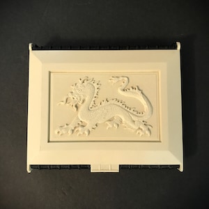 May include: A white decorative box with a raised image of a dragon on the front. The box has a black trim around the edges.