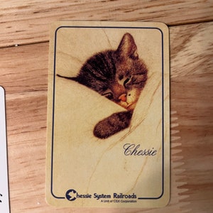 K&ouml;nnte beinhalten: Eine Karte im Vintage-Stil mit einem schlafenden Tabby-Kater, dessen Name "Chessie" in Kursivschrift geschrieben ist. Die Karte hat einen braunen Hintergrund mit einem blauen Rand und den Text "Chessie System Railroads, A Unit of CSX Corporation" unten.
