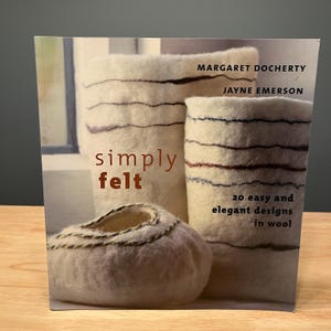 May include: A softcover book titled Simply Felt by Margaret Docherty and Jayne Emerson, featuring 20 easy and elegant designs in wool. The cover displays three off-white felted vessels with dark horizontal stitching details.