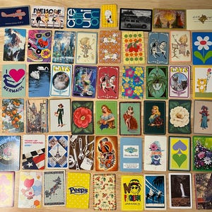 May include: A collection of 48 playing cards with various designs and themes. The cards feature images of mermaids, cats, flowers, airplanes, and more. The cards are arranged in a grid pattern on a wooden surface.