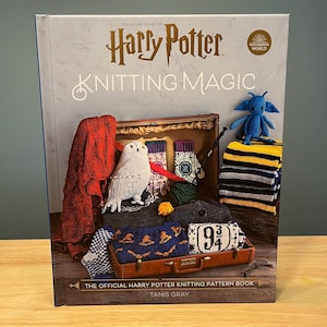 May include: A hardcover book titled "Harry Potter Knitting Magic" with a suitcase filled with knitted items, including a red scarf, owl, gloves, and a blue creature. The book cover features the Harry Potter logo and the Wizarding World logo.