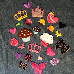 May include: Assortment of colorful iron-on patches featuring various designs, including cupcakes, hearts, crowns, butterflies, a castle, and a bow. The patches are perfect for adding a touch of whimsy to clothing, bags, or other fabric items.