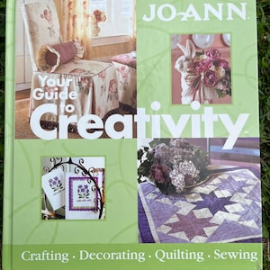 May include: A hardcover book titled JO-ANN Your Guide to Creativity with a light green cover. The front features images of a floral chair, a rabbit with flowers, embroidered cards, and a purple star-patterned quilt. Text includes Crafting, Decorating, Quilting, Sewing.