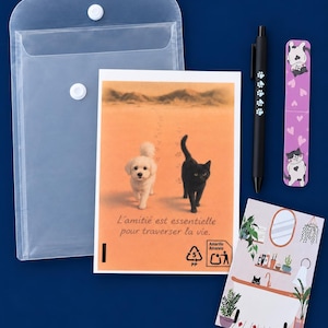 Puede incluir: Un sobre de plástico translúcido, un bolígrafo negro con huellas blancas, un marcapáginas rosa con ilustraciones de gatos y dos tarjetas rectangulares. Una tarjeta muestra un perro blanco y un gato negro con el texto L'amitié est essentielle pour traverser la vie.