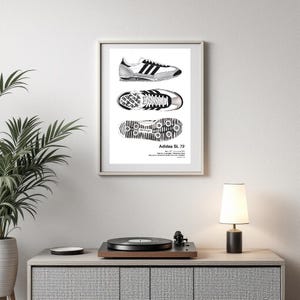 May include: Framed black and white print of Adidas SL 72 trainers, showing top, bottom, and side views. The trainers have black stripes and a white sole. The print is displayed on a white wall.