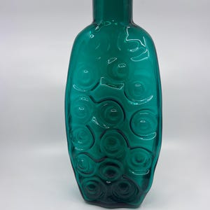 Pode incluir: Vaso de vidro verde-azulado com uma superfície texturizada e um padrão de círculos giratórios. O vaso tem um pescoço alto e fino e um corpo arredondado. O vidro é transparente.