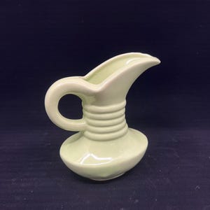 May include: A small, pale green ceramic jug with a curved spout and a rounded handle. The jug has a bulbous base and a ridged neck, giving it a vintage or retro aesthetic. The background is a dark, solid colour.