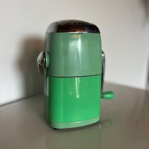 May include: A retro ice crusher in mint green with a chrome top and accents. The hand-cranked appliance has a cylindrical shape, a small green handle, and a silver crank. This vintage kitchen tool is designed for crushing ice.