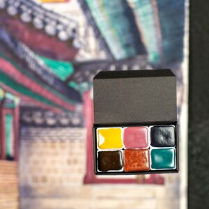 Könnte beinhalten: Ein Aquarellgemälde eines Gebäudes mit roten, grünen und blauen Akzenten. Eine kleine schwarze Box mit Aquarellfarben in Gelb, Rosa, Braun, Orange und Teal liegt vor dem Gemälde.