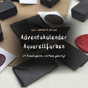 Könnte beinhalten: Eine Sammlung von vierundzwanzig kleinen, schwarz verpackten Aquarellfarben-Näpfchen, einige mit Farbmustern auf weißem Papier, mit dem Text 2026 - LIMITIERTE AUFLAGE, Adventskalender Aquarellfarben, 24 Einzelpigmente, von Hand gefertigt.
