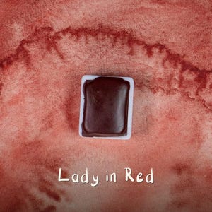 Könnte beinhalten: Ein rechteckiges, dunkelbraunes Aquarellfarben-Pfännchen in einem weißen Kunststoffbehälter, zentriert vor einem strukturierten, roten Aquarell-Hintergrund. Der Text "Lady in Red" ist in einer einfachen weißen Schrift unten abgebildet.