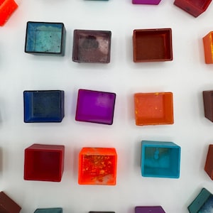 Könnte beinhalten: Draufsicht auf eine Sammlung kleiner, quadratischer Aquarellfarben-Näpfchen in verschiedenen Farben. Die Näpfchen sind auf einer weißen Oberfläche angeordnet und zeigen Farbtöne von Blau, Lila, Orange und Rot. Die transparente Natur der Farbe ist sichtbar.