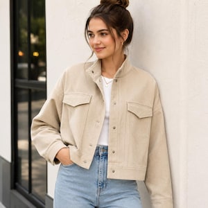 May include: A beige corduroy jacket featuring a structured collar, long sleeves, and two chest pockets with flaps. The jacket has a button-front closure with visible metallic buttons and a cropped hemline, worn over a plain white shirt and blue jeans.