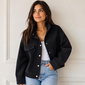 May include: A black corduroy jacket featuring a structured collar, long sleeves, and a front button-down closure with silver-toned metal buttons. The jacket includes a single chest pocket with a flap and a straight hemline, worn over a white top.