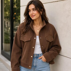 May include: A brown corduroy jacket featuring a collared neckline, long sleeves, and a button-down front closure. The jacket includes two chest pockets with flaps and is worn over a plain white top paired with light blue denim jeans.