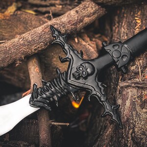 K&ouml;nnte beinhalten: Schwarzes Fantasy-Schwert mit Totenkopf-Design am Parierst&uuml;ck und wei&szlig;er Klinge. Das Schwert liegt auf einem Bett aus Holzscheiten und schafft eine rustikale und thematische Umgebung.