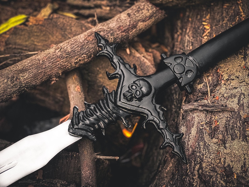 K&ouml;nnte beinhalten: Schwarzes Fantasy-Schwert mit Totenkopf-Design am Parierst&uuml;ck und wei&szlig;er Klinge. Das Schwert liegt auf einem Bett aus Holzscheiten und schafft eine rustikale und thematische Umgebung.