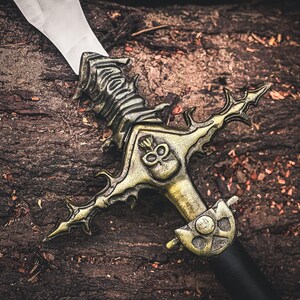 K&ouml;nnte beinhalten: Ein dekoratives Schwert mit einer silbernen Klinge und einem verzierten, goldfarbenen Knauf. Der Knauf weist ein Totenkopf-Design und aufwendige Details auf. Das Schwert liegt auf einer strukturierten, braunen Oberfl&auml;che, was ein Fantasy- oder historisches Thema suggeriert.