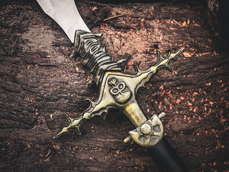 K&ouml;nnte beinhalten: Ein dekoratives Schwert mit einer silbernen Klinge und einem verzierten, goldfarbenen Knauf. Der Knauf weist ein Totenkopf-Design und aufwendige Details auf. Das Schwert liegt auf einer strukturierten, braunen Oberfl&auml;che, was ein Fantasy- oder historisches Thema suggeriert.