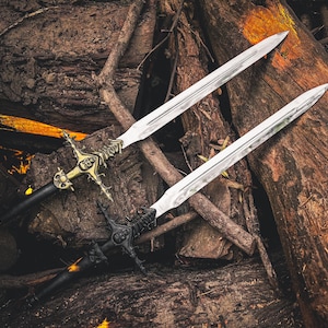 K&ouml;nnte beinhalten: Zwei dekorative Schwerter mit verzierten Griffen und silbernen Klingen liegen auf einem Holzbett. Ein Schwert hat einen goldfarbenen Griff mit aufwendigen Details, das andere einen schwarzen Griff. Die Klingen sind lang und spitz.