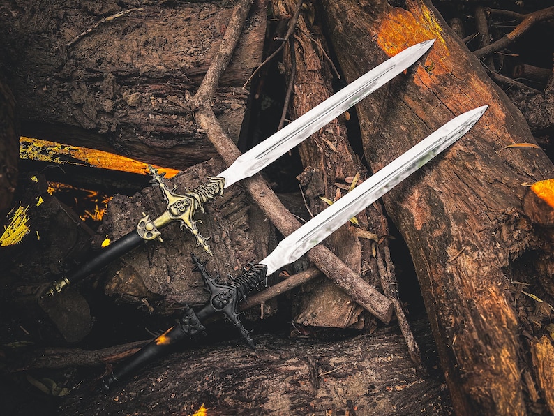 K&ouml;nnte beinhalten: Zwei dekorative Schwerter mit verzierten Griffen und silbernen Klingen liegen auf einem Holzbett. Ein Schwert hat einen goldfarbenen Griff mit aufwendigen Details, das andere einen schwarzen Griff. Die Klingen sind lang und spitz.