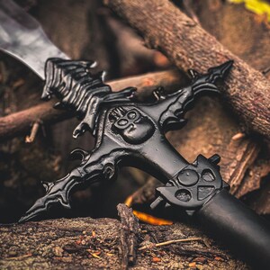 K&ouml;nnte beinhalten: Schwarzes Fantasy-Schwert mit aufwendigen Details, darunter ein Totenkopf-Design am Parier. Die Klinge ist silberfarben, der Griff schwarz. Das Schwert liegt auf Holz und &Auml;sten.