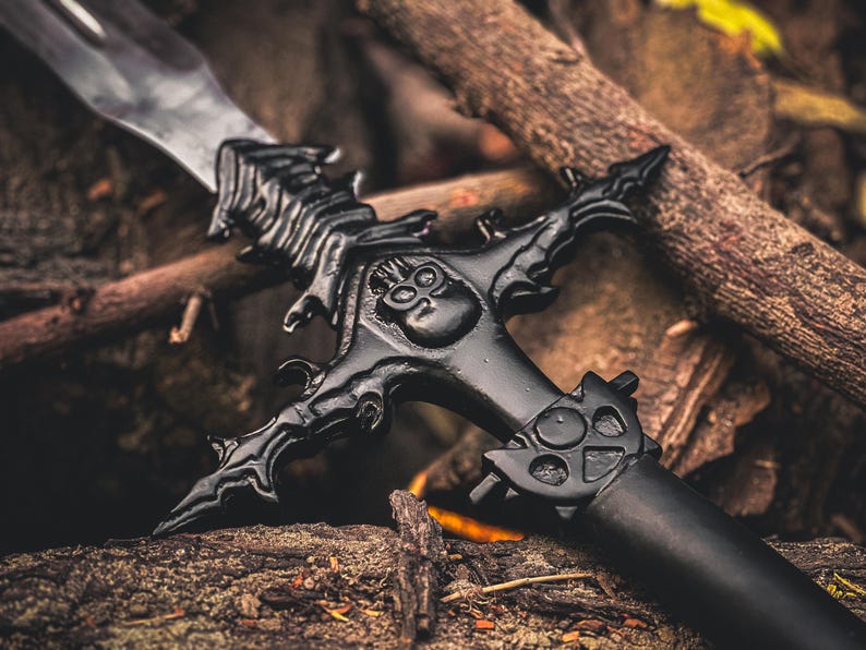 K&ouml;nnte beinhalten: Schwarzes Fantasy-Schwert mit aufwendigen Details, darunter ein Totenkopf-Design am Parier. Die Klinge ist silberfarben, der Griff schwarz. Das Schwert liegt auf Holz und &Auml;sten.