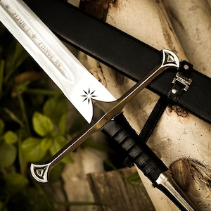 Puede incluir: Espada plateada y dorada con una vaina de cuero negro. La hoja de la espada tiene texto grabado y un dise&ntilde;o de estrella. La empu&ntilde;adura es dorada con detalles plateados. La vaina es negra con herrajes plateados.